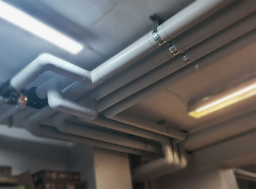 Insulated plumbing pipes covered with Isover insulation and Isogen protective cladding, installed under concrete ceiling in a technical room.